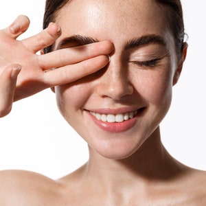 May include: A close-up of a woman's face with freckles and a bright smile. Her eyes are closed and her hand is covering one eye. Her skin is clear and glowing.