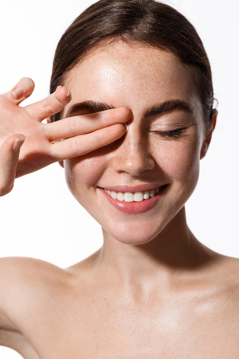 May include: A close-up of a woman's face with freckles and a bright smile. Her eyes are closed and her hand is covering one eye. Her skin is clear and glowing.
