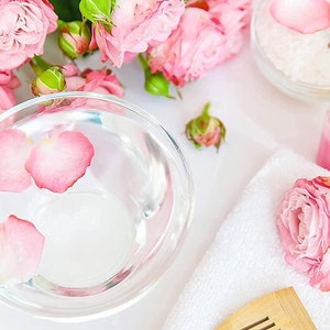 May include: A white towel with a wooden comb, a bowl of water with pink rose petals, a small bottle of pink liquid, a small bottle of yellow liquid, and pink roses.