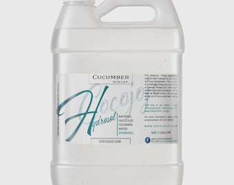 Puede incluir: Una garrafa de plástico transparente de un galón que contiene hidrosol de pepino. La etiqueta muestra el texto 'CUCUMBER', 'COCOJOJO' y 'NATURAL DISTILLED CUCUMBER WATER HYDROSOL'. Incluye un tapón de rosca blanco y un asa integrada en el diseño.