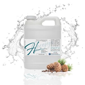 May include: A clear plastic jug of Cedarwood Hydrosol, a 100% natural, pure distilled floral water hydrosol. The jug is sitting on a white surface with water splashing around it. The label on the jug says "Cocojojo" and "Cedarwood Hydrosol". There are pine cones and needles in the bottom right corner of the image.