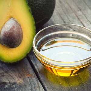 May include: A small glass bowl filled with golden yellow avocado oil sits on a rustic wooden surface next to a halved avocado.