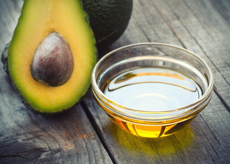 May include: A small glass bowl filled with golden yellow avocado oil sits on a rustic wooden surface next to a halved avocado.