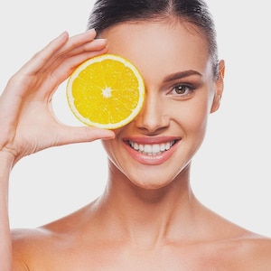 May include: A woman holds a bright yellow orange slice over one eye, smiling. The orange slice is round with a visible center. The woman has brown hair and is wearing no top. The background is a solid white.