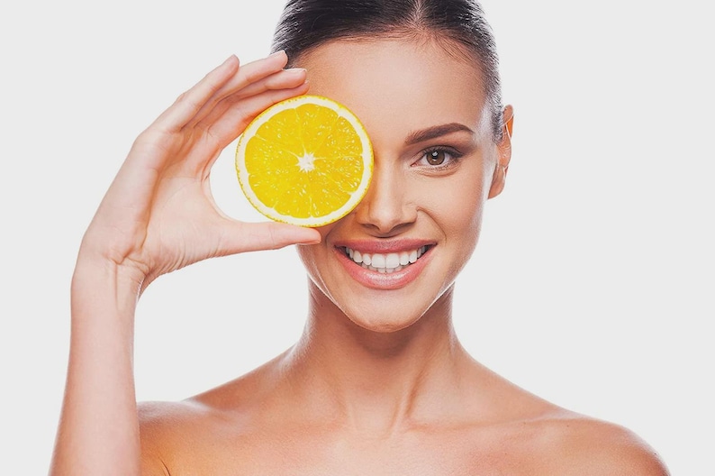 May include: A woman holds a bright yellow orange slice over one eye, smiling. The orange slice is round with a visible center. The woman has brown hair and is wearing no top. The background is a solid white.