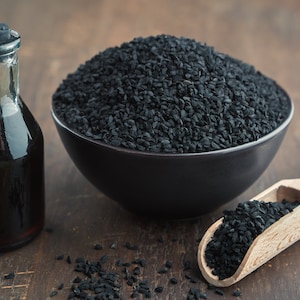 K&ouml;nnte beinhalten: Eine schwarze Sch&uuml;ssel gef&uuml;llt mit schwarzen Kreuzk&uuml;mmelsamen, eine kleine Flasche mit schwarzem Kreuzk&uuml;mmel&ouml;l und eine Holzschaufel mit schwarzen Kreuzk&uuml;mmelsamen.