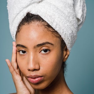 May include: A woman with dark brown skin is wearing a white towel wrapped around her head. She is looking at the camera with a serious expression. Her hand is resting on her cheek.