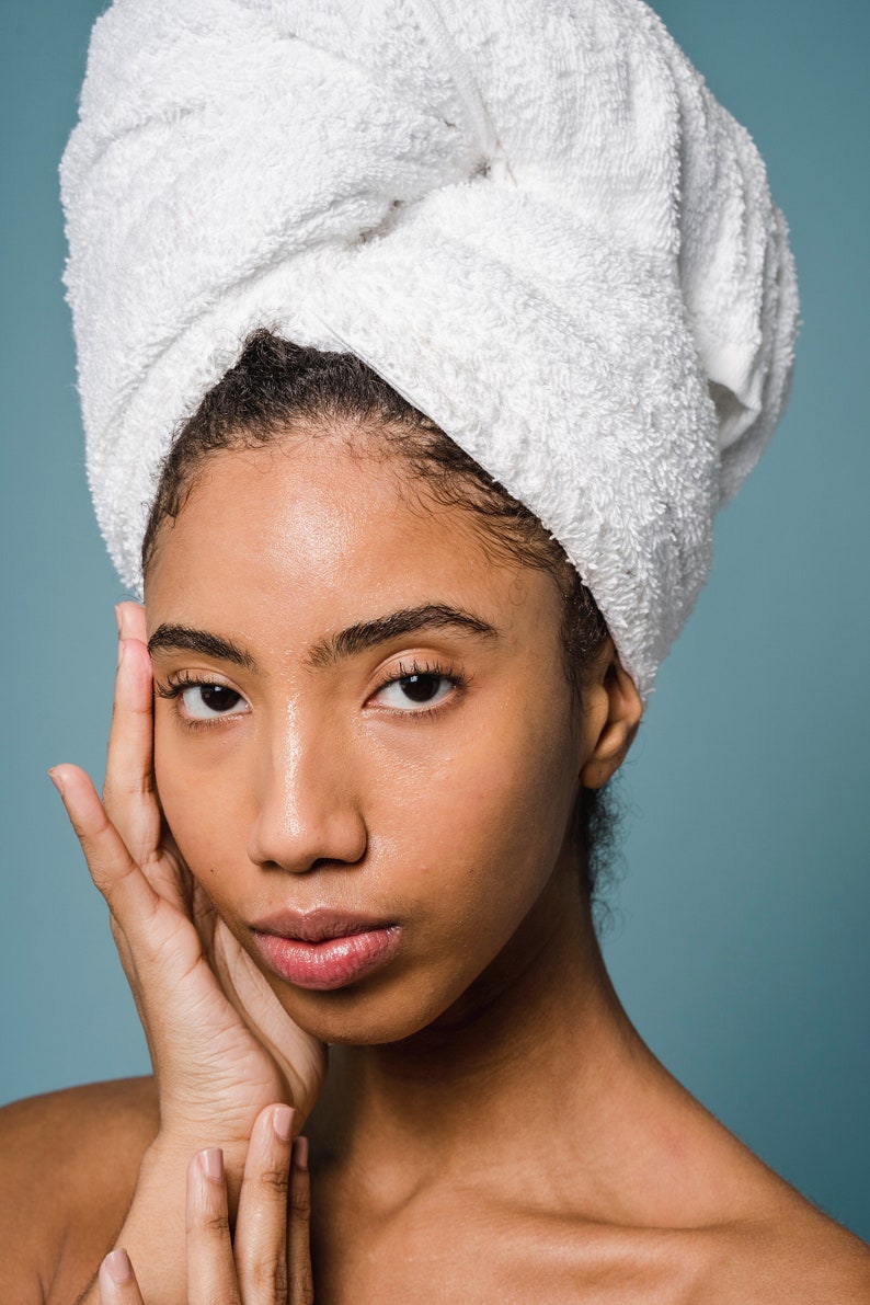May include: A woman with dark brown skin is wearing a white towel wrapped around her head. She is looking at the camera with a serious expression. Her hand is resting on her cheek.