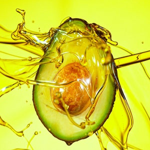 May include: A halved avocado is suspended in a splash of yellow oil, creating a dynamic and colorful image.