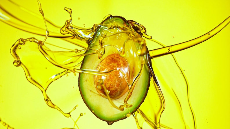 May include: A halved avocado is suspended in a splash of yellow oil, creating a dynamic and colorful image.