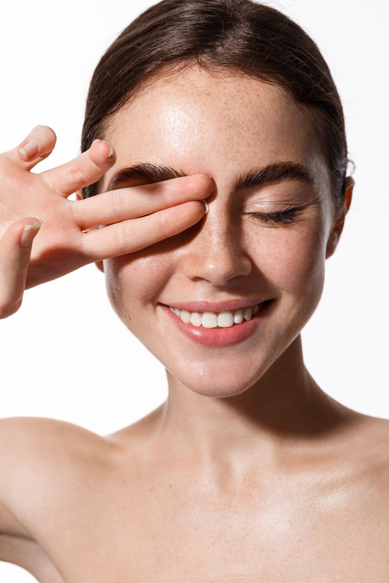 May include: A close-up of a woman's face with freckles and a bright smile. Her eyes are closed and her hand is covering one eye. Her skin is clear and glowing.