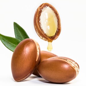 May include: Close-up of three brown argan nuts with one nut split open, revealing the white kernel and a drop of golden oil dripping from it. Green leaves are in the background.