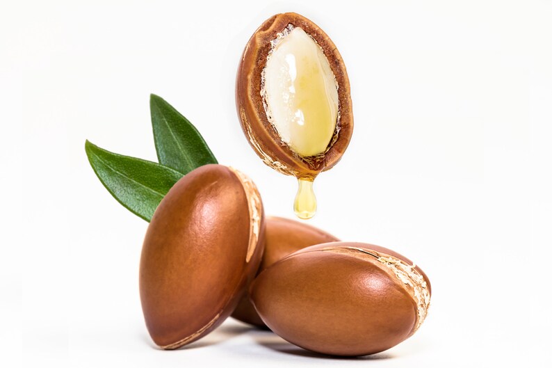 May include: Close-up of three brown argan nuts with one nut split open, revealing the white kernel and a drop of golden oil dripping from it. Green leaves are in the background.