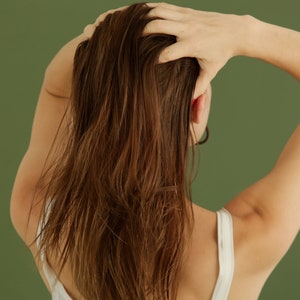 May include: A person with long brown hair is shown from behind, with their hand reaching up to touch their hair. They are wearing a white tank top.