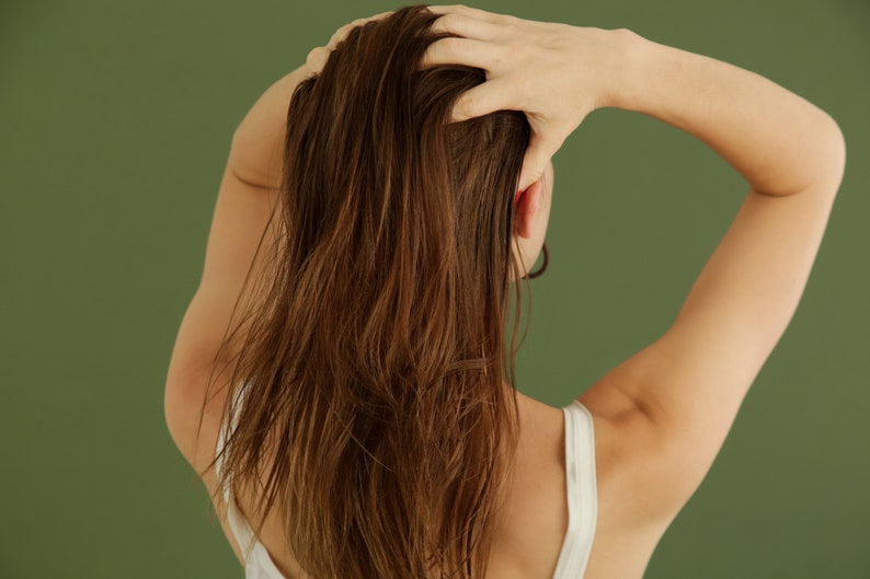 May include: A person with long brown hair is shown from behind, with their hand reaching up to touch their hair. They are wearing a white tank top.