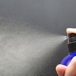 May include: A hand holding a blue glass spray bottle with a black nozzle, spraying a fine mist into the air.