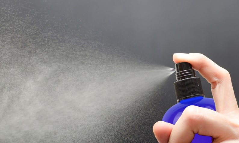 May include: A hand holding a blue glass spray bottle with a black nozzle, spraying a fine mist into the air.
