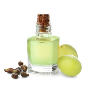 May include: A clear glass bottle with a cork stopper, filled with a pale yellow liquid. Two green grapes and a pile of grape seeds are next to the bottle. The background is white.