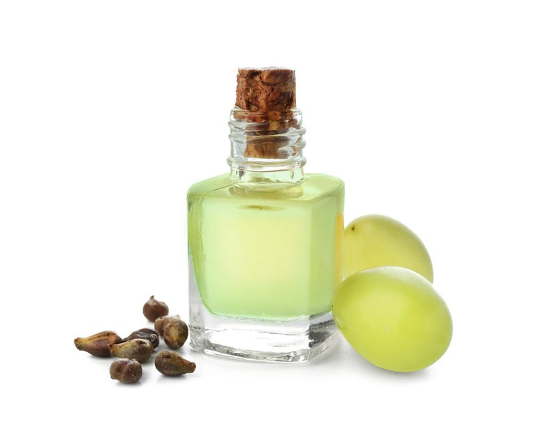 May include: A clear glass bottle with a cork stopper, filled with a pale yellow liquid. Two green grapes and a pile of grape seeds are next to the bottle. The background is white.