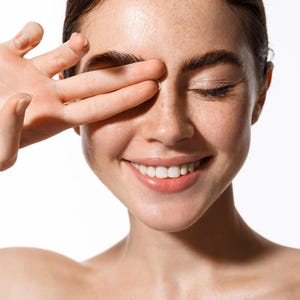 May include: A smiling person with freckles, eyes closed, and a hand covering one eye. The person has dark brown hair and is looking towards the right. The background is white.
