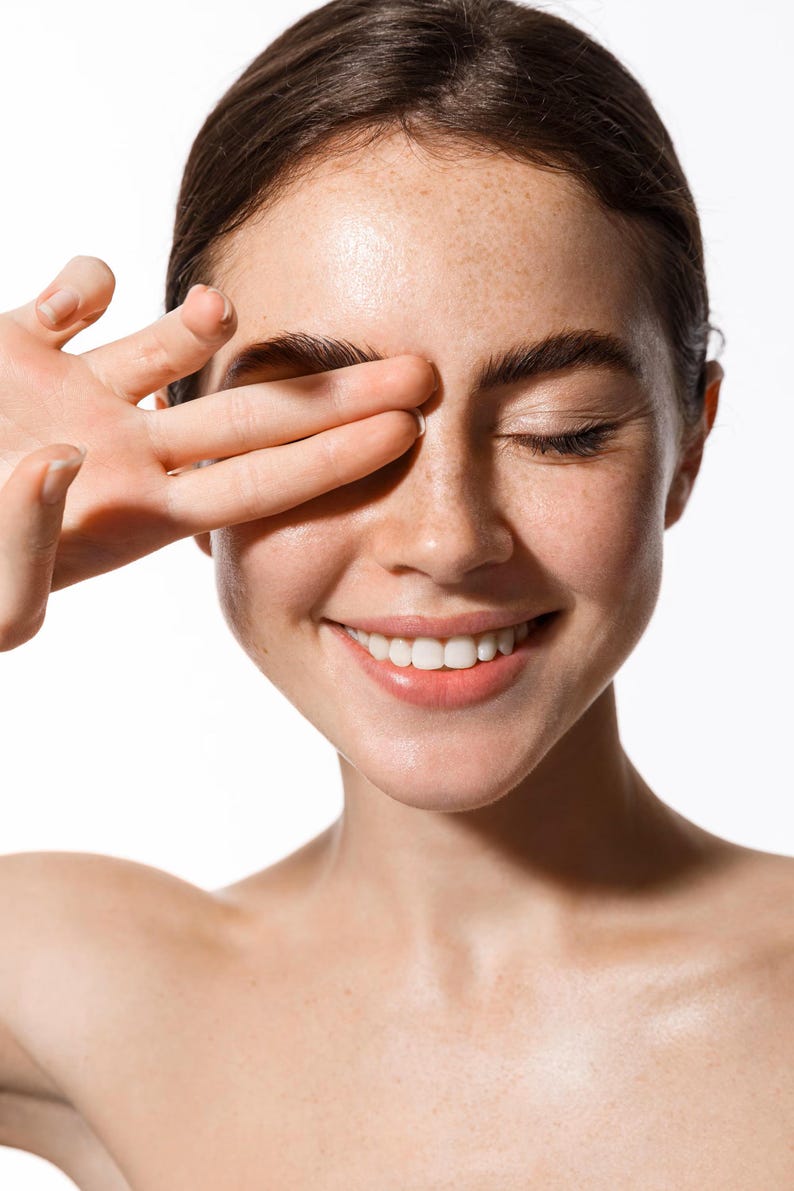 May include: A smiling person with freckles, eyes closed, and a hand covering one eye. The person has dark brown hair and is looking towards the right. The background is white.