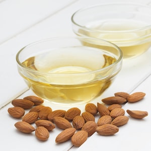 Puede incluir: Dos cuencos de vidrio transparente llenos de aceite de almendras amarillo claro, rodeados de una dispersi&oacute;n de almendras marrones sobre una superficie de madera blanca.