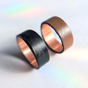 May include: Two rings with a smooth, metallic copper-coloured interior band. One ring features a textured black exterior surface, while the other ring features a textured tan-coloured exterior surface. Both rings have a wide, flat band design.
