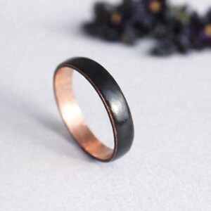 May include: A ring with a smooth, matte black exterior surface and a polished, metallic copper-coloured interior band. The ring features a simple, rounded profile and is positioned upright to display both the dark outer finish and the warm inner tone.