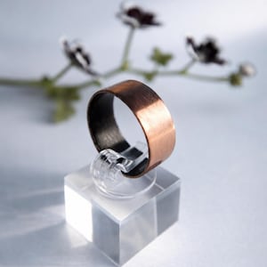 May include: A wide band ring featuring a brushed copper exterior finish and a dark, matte black interior surface. The ring is displayed upright on a clear, square acrylic stand, showcasing its metallic texture and contrasting colour scheme.