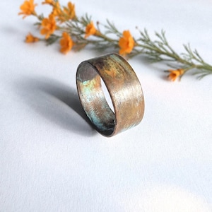 Peut inclure: Une bague large à la finition métallique patinée, présentant un mélange de tons cuivre, brun et vert sarcelle oxydé. La surface offre un aspect texturé et irrégulier avec des coups de pinceau visibles et une patine rustique vieillie.