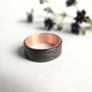 Peut inclure: Une bague dotée d'une bande extérieure gris anthracite texturée et sombre, contrastant avec un intérieur lisse et poli aux tons cuivrés. L'anneau présente un profil plat et une largeur constante, mettant en valeur le contraste des matériaux.