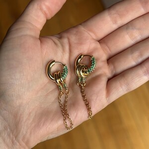 Op de afbeelding: Een paar goudkleurige oorringen met een scharniersluiting. Elke ring is versierd met een segment van kleine, levendige groene steentjes. Aan elke ring hangt een fijn goudkleurig kettinkje, wat zorgt voor een bungelend effect.