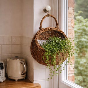 May include: A round, brown wicker basket with a handle, hanging on a wall. The basket is filled with green plants with trailing vines. The basket is suspended from a metal hook attached to a white wall.