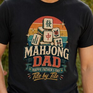 May include: A black t-shirt featuring a graphic of stacked mahjong tiles in front of a retro-style sunset. The text reads "MAHJONG DAD", "HAPPY FATHER'S DAY", and "Tile by Tile" in a vintage font. The design includes stars and decorative flourishes.