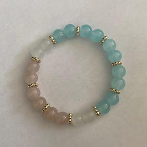 Op de afbeelding: Een armband met ronde kralen in lichtblauw, roze en wit. Goudkleurige afstandhouders scheiden de kralen. De armband is ontworpen om om de pols te worden gedragen.