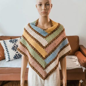 Puede incluir: Un poncho de ganchillo hecho a mano y colorido con un patrón geométrico. El poncho presenta rayas marrones, amarillas, rosas, verdes, azules y blancas. El cuello y los bordes están ribeteados con hilo marrón más oscuro. El poncho se exhibe en un maniquí.