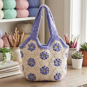 Könnte beinhalten: Eine handgefertigte Häkeltasche mit floralem Design. Die Tasche hat eine cremefarbene Basis mit sechseckigen Paneelen, die jeweils mit einer blauen Blume und einem gelben Zentrum verziert sind. Oberteil und Griffe sind in passendem Blau gehalten.