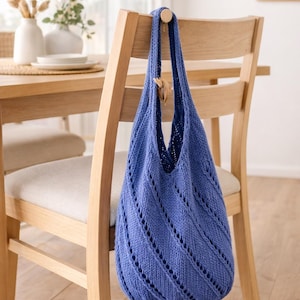May include: A blue knitted tote bag with a diagonal openwork pattern hangs on a wooden chair. The bag has a wide opening and a rounded bottom. The chair is light-colored wood, and the background includes a table and decorative items.