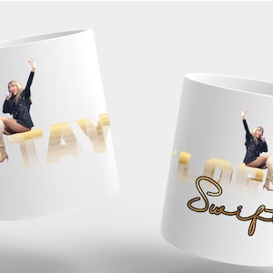Puede incluir: Dos tazas de cer&aacute;mica blanca con asa. Una taza tiene la palabra "TAY" en letras doradas con la imagen de una persona. La otra taza tiene la palabra "Swift" en letras doradas con la imagen de una persona.
