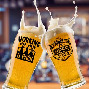 May include: Two clear glass beer glasses filled with golden lager, each featuring black printed text and graphics. The left glass displays 'WORKING on my 6 PACK' with bottle icons, while the right glass shows 'IT'S BEER O'CLOCK' with a beer mug icon.