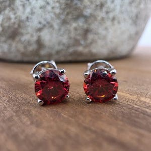 May include: A pair of stud earrings featuring round, faceted, deep red stones set in four-prong silver-toned metal settings. The earrings are positioned side by side, showcasing the vibrant red color and the reflective quality of the cut stones.