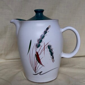May include: A white ceramic teapot with a dark teal lid and a handle on the right side. The body features a painted design of stylized wheat stalks in shades of teal, red, and black, with a curved black line accent at the base of the illustration.