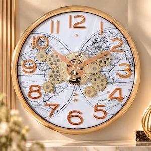 May include: A round wall clock with a gold frame and a white face featuring a black world map print. The clock has raised gold-tone numerals, gold-tone hands, and a central exposed gear mechanism with multiple small gold-tone gears.