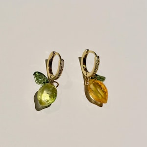 Peut inclure: Une paire de boucles d'oreilles créoles dorées. Chaque boucle d'oreille présente un petit charme de citron translucide avec une feuille verte et un cerceau doré avec de petits cristaux transparents. Un citron est jaune-vert, l'autre est jaune-orange.