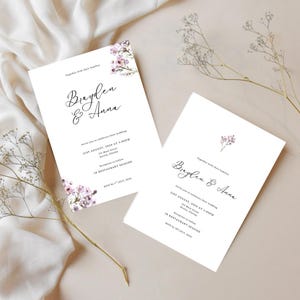May include: Two white wedding invitations with floral accents and elegant script. The invitations read "Braylen & Anna" and include event details. Delicate sprigs of flowers are arranged around the invitations.