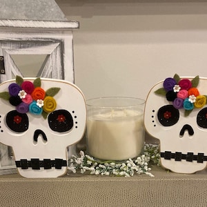 May include: Two white wooden sugar skull decorations with black eyes and mouths. Each skull has a colourful felt flower crown with green leaves.