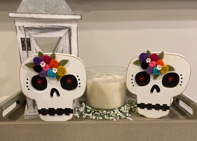 May include: Two white wooden sugar skull decorations with black eyes and mouths. Each skull has a colourful felt flower crown with green leaves.