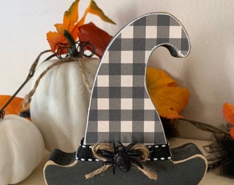 May include: A wooden witch hat decoration with a black and white checkered pattern. The hat has a black base and a black ribbon with a glittery spider and twine bow. The background includes white pumpkins and fall foliage.