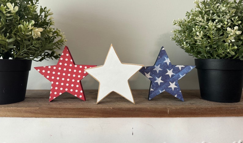 May include: Three wooden stars, each with a different design. The first star is red with white polka dots, the second star is white with a gold outline, and the third star is blue with white stars.