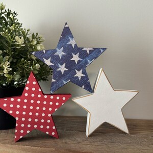 May include: Three wooden stars in red, white, and blue patriotic colors. The red star has white polka dots, the blue star has white stars, and the white star has a gold outline.